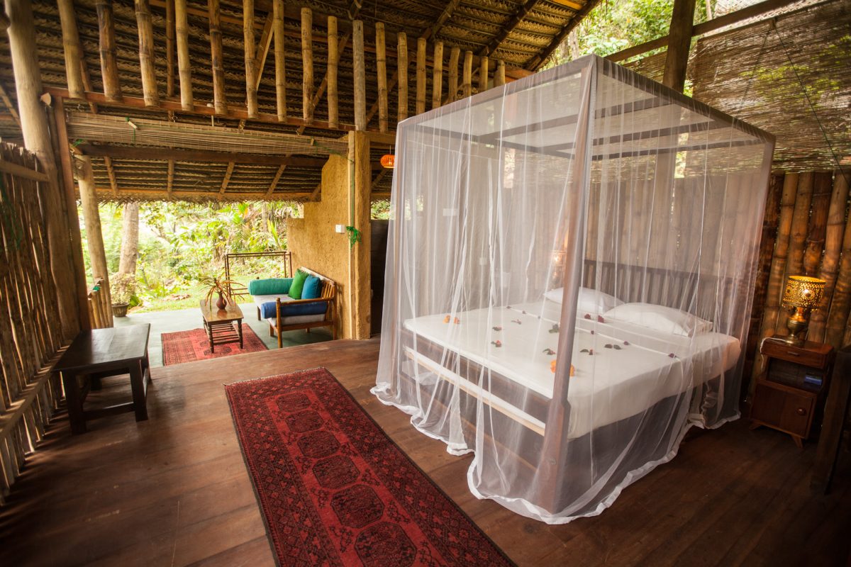 Bamboo hut interior with a four-poster bed and daybed opening to the garden at Talalla Retreat, Sri Lanka